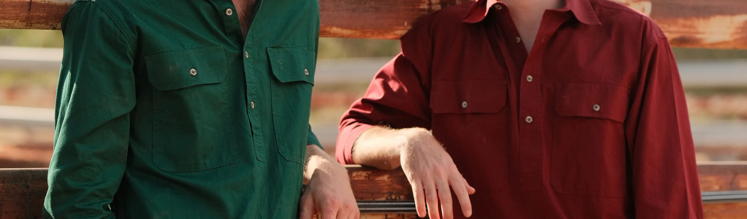 Two men in green and red closed-front work shirts lean on a stockyard rail